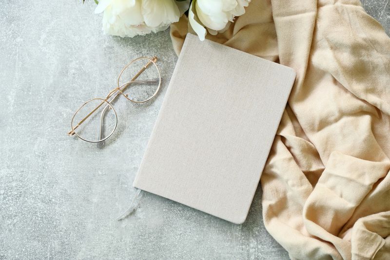 Flat lay paper notebook, eyeglasses, peony flowers and beige cloth on stone background. Top view girl home office desk with elegant accessories. Modern feminine workspace concept