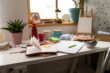 A cluttered white desk with art supplies and plants near a window.