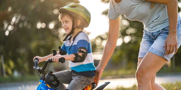 A young boy learns to ride a bike with a woman’s support outdoors.