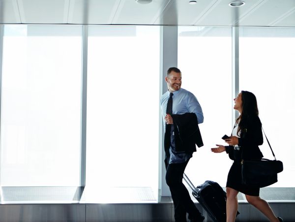 Two business travelers walk and chat with luggage in an airport.