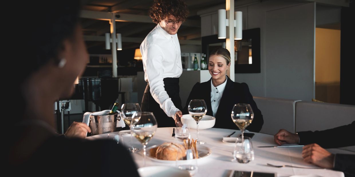 A waiter serves food to a smiling woman at a fine dining restaurant.
