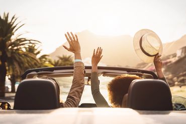 Two friends enjoying a sunny drive in a convertible car with hands raised.