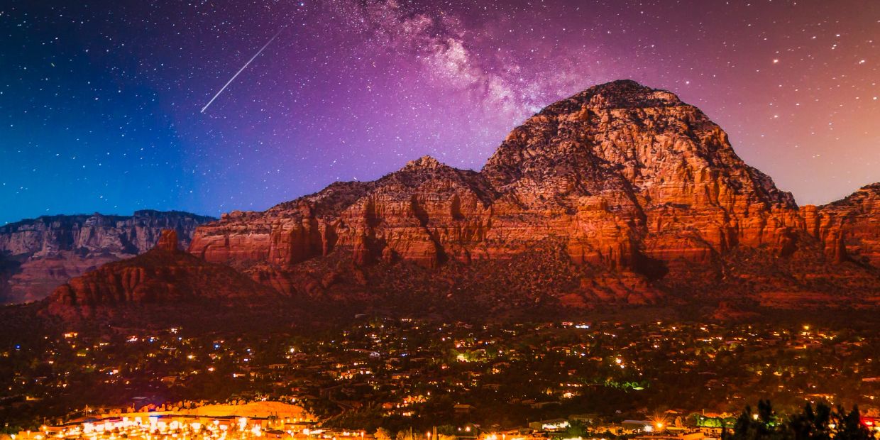 Night sky with shooting star over illuminated red rock mountains.