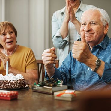 Elderly man celebrating birthday with thumbs up and smiling companions.