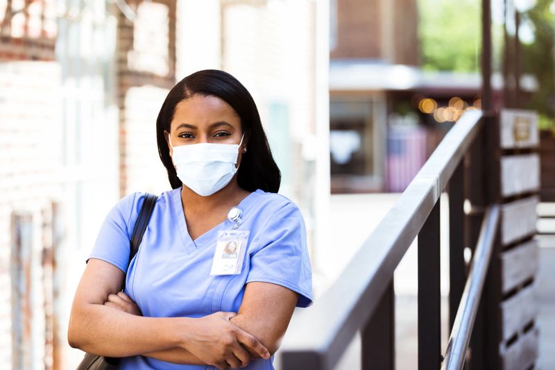 A mid adult African American nurse looks at the camera while standing with her arms crossed. She is wearing a protective face mask, as she prepares for her shift in the COVID-19 ward.