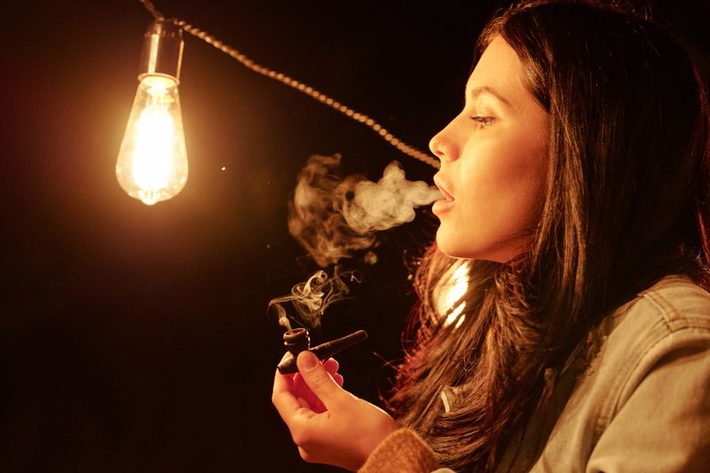 Young woman with dark long hair holding wooden tobacco pipe while smoking at night in the street and breathing smoke out of mouth