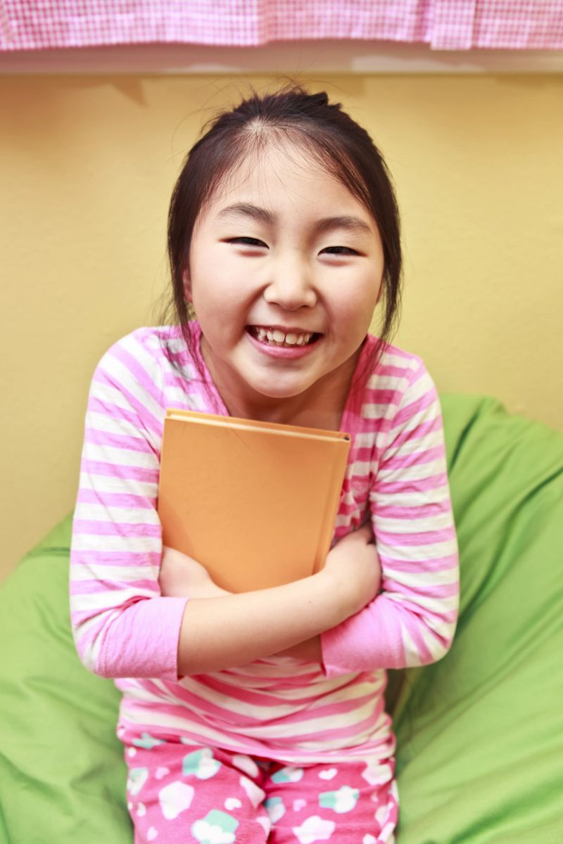 7 years old cute Asian girl in pajama, sitting on a bean bag, hugging a book tightly in her arms and smiling at the camera.
