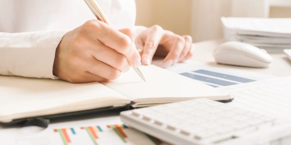 Person writing notes at a desk with financial charts and calculator.