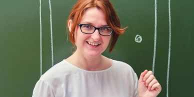 Smiling woman with red hair and glasses in front of a chalkboard.