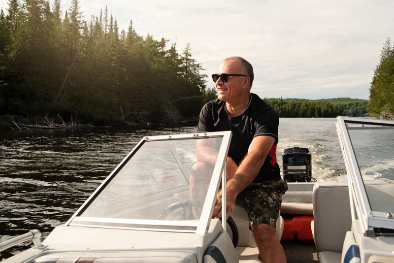 50 + man driving small boat on a lake. He is wearing hat and  black t-shirt over swimwear, and is looking at the horizon. Horizontal waist up outdoors shot with copy space.