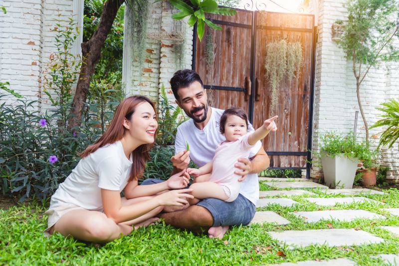 Mixed race family concept. Happy father mother and little infant baby daughter sitting on garden at house. Parent and adorable baby girl get relax and happy with smile face, daughter point finger