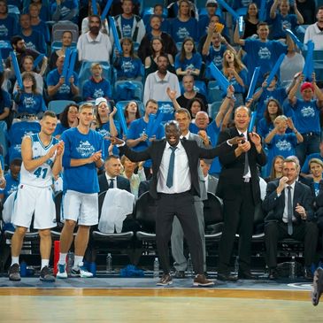Basketball team and coach celebrate enthusiastically in front of cheering fans.