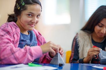 Two girls engaged in crafting paper pyramids at a table.
