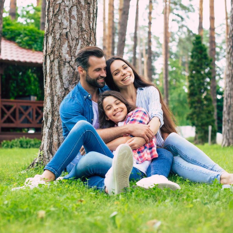 Beautiful happy family while sitting together on the grass and hugging each other, relaxing outdoors on suburban house background.