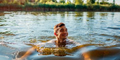 A smiling young man swimming in a calm lake surrounded by greenery.