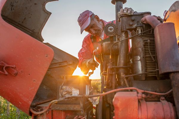 An older man repairs a tractor engine at sunset.