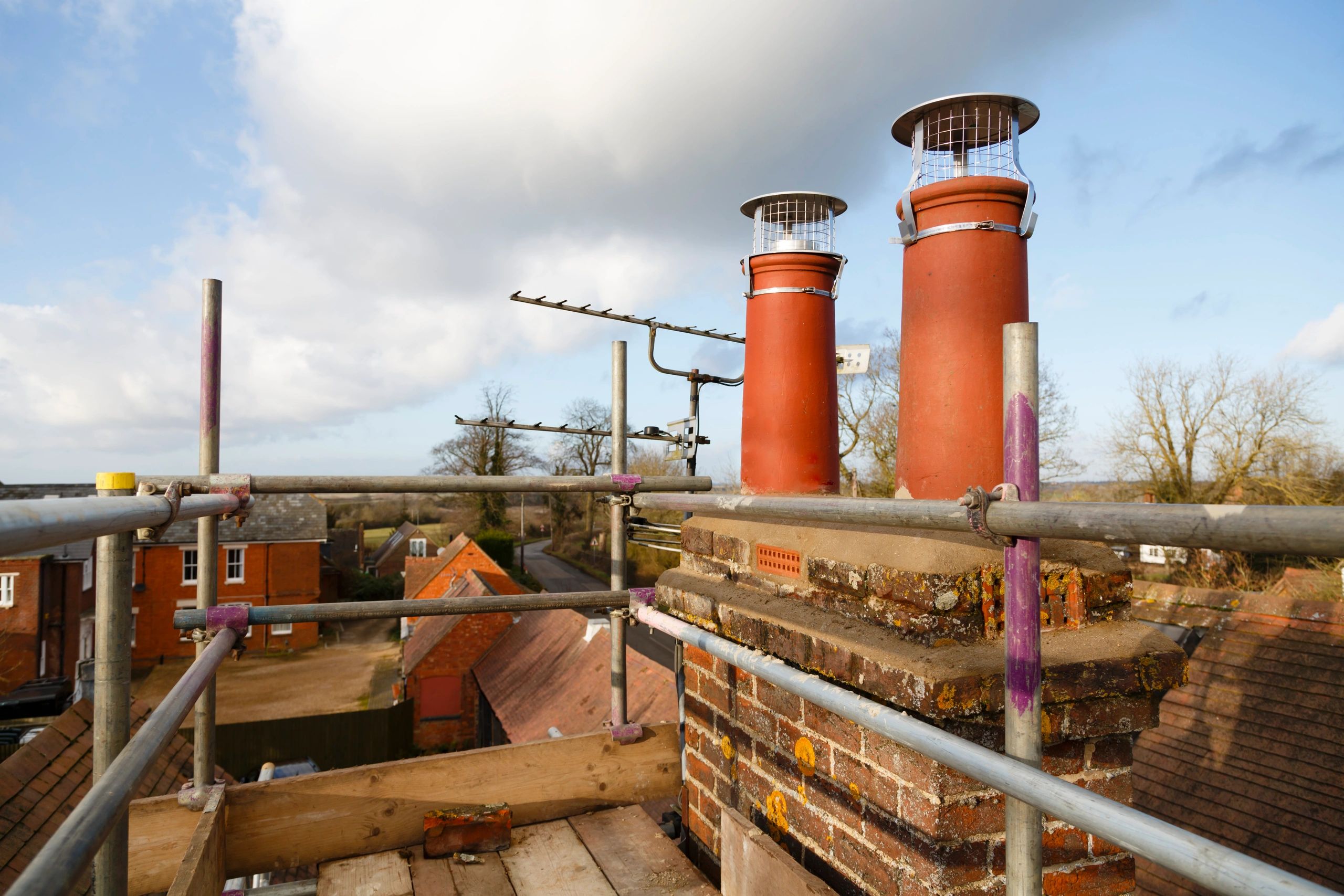 View of two red chimney pots on a brick chimney with scaffolding and rooftops below.