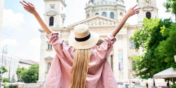 Woman with long hair and hat raising arms in front of historic cathedral.