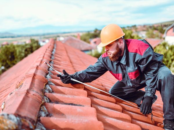 A roofer measures red tiles on a house roof wearing safety gear.