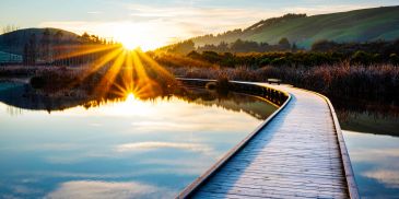 Sunset over a serene lake reflecting the sky, with a wooden path and green hills.
