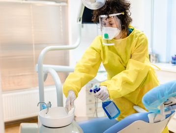 Healthcare worker disinfecting a dental chair area wearing protective gear.