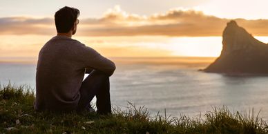A man sitting on grass, gazing at a peaceful sunset over the ocean with a distant rocky island.