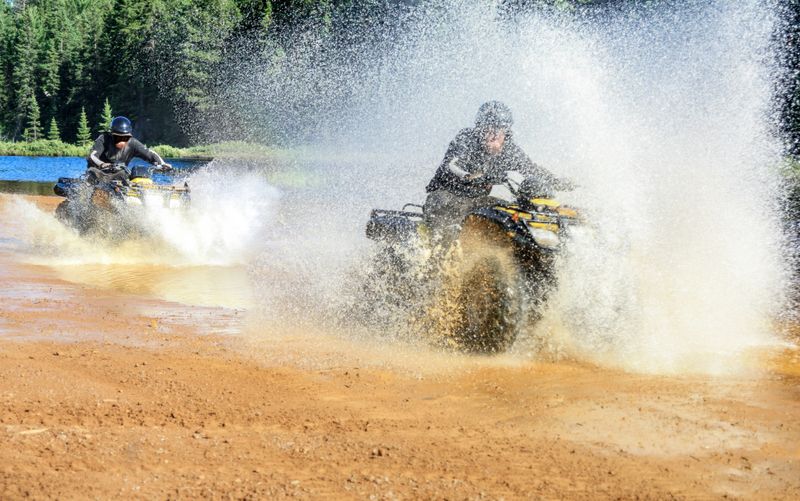 Two Men having Fun and Enjoy driving ATVs quads through splashing lake water with high speed on a beach of Foyross Lake. Foy, Sudbury, Canada.
