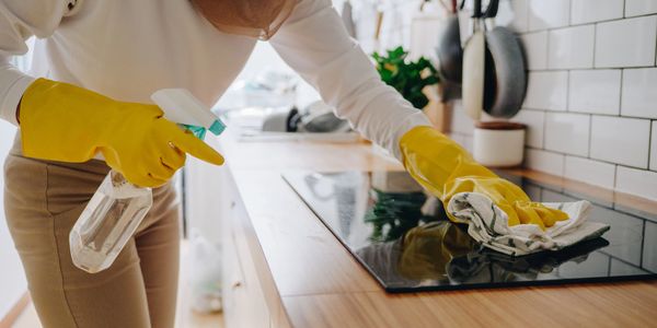 Person cleaning kitchen stove with yellow gloves and spray bottle.