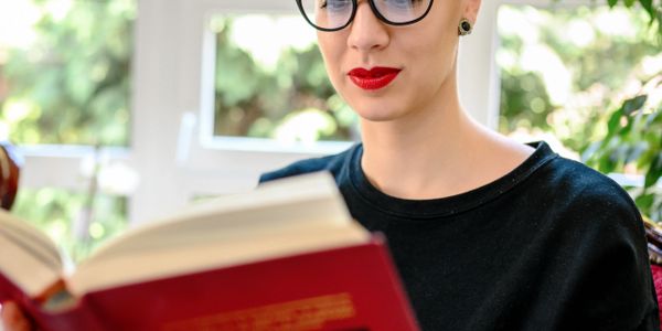 Person with glasses and red lipstick reading a book indoors.