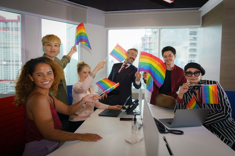 diverse group of business people (man, woman, gay, transgender, lesbian, asian, caucasian, african american, lgbtq) with rainbow flag on hand in business office working together as teamwork