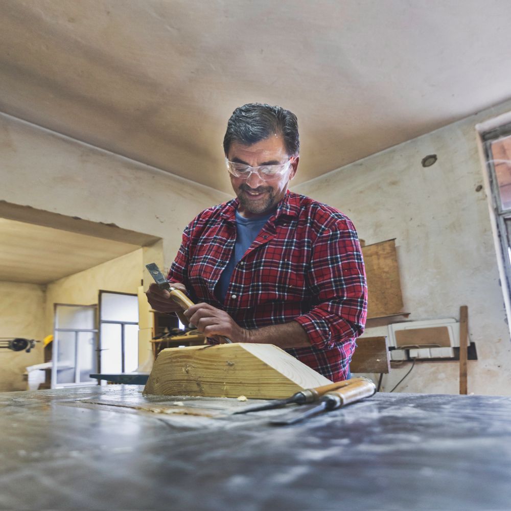 Man wearing safety goggles carves wood in a workshop.