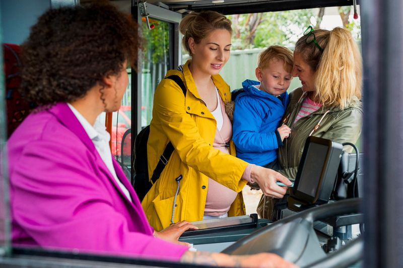 Mature caucasian woman, her pregnant daughter, and grandson getting onto a bus shot through a window over the mixed-race female bus driver's shoulder.