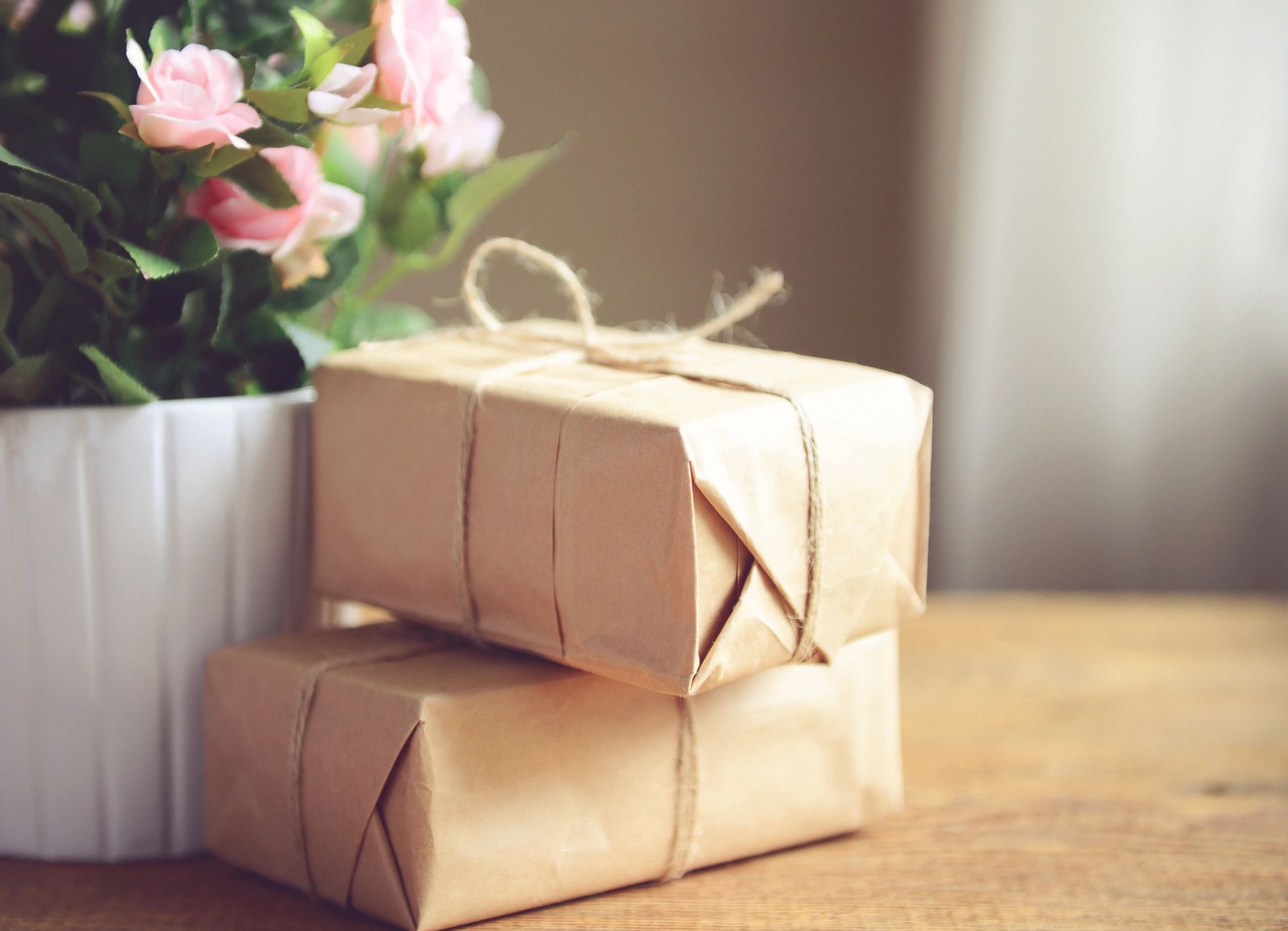Two kraft-paper wrapped gifts stacked by a white vase filled with blush flowers on a wood table.