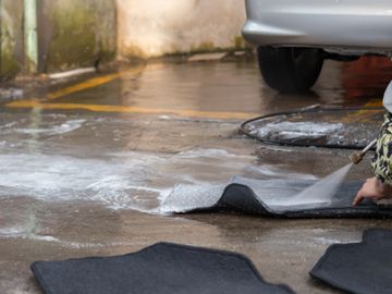 Person washing car floor mats with water spray near a silver car.