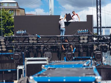 Two workers install a large LED screen on an outdoor stage setup.
