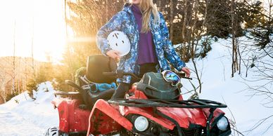 Woman with white helmet standing on red ATV in snowy forest during sunset.