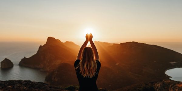 Person holding the sun at sunset over a mountainous coastline.