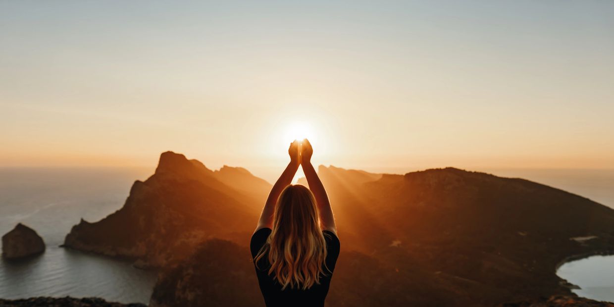 Woman holding the sun at sunset over mountains and sea.