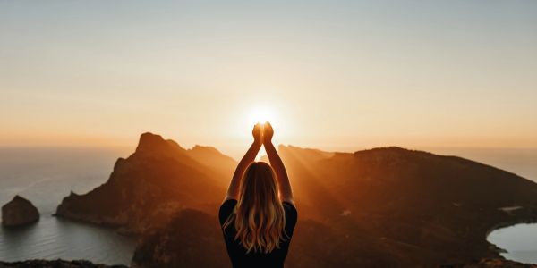 Person holding the sun at sunset over mountains.
