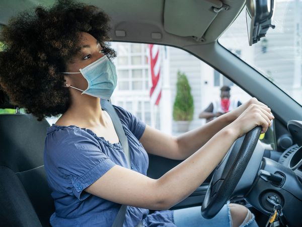 Woman wearing a mask driving a car with an American flag outside.
