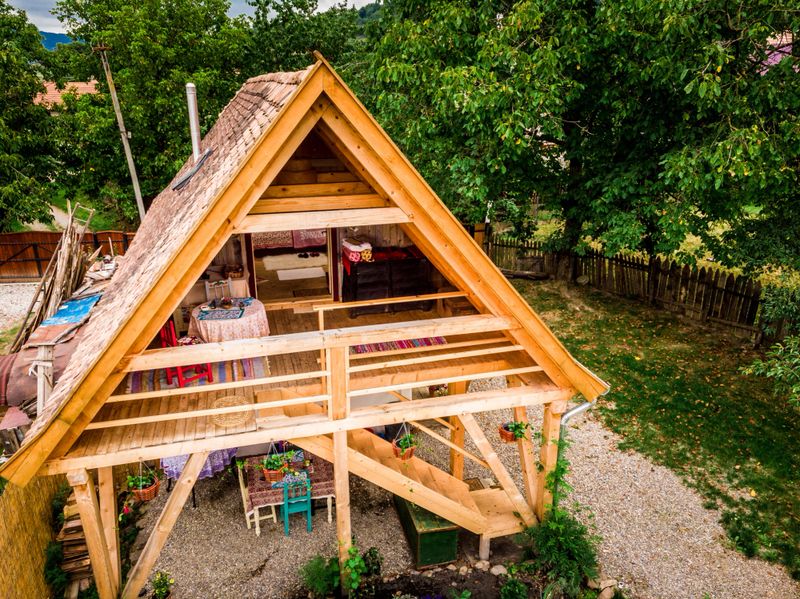 Aerial drone shot depicting a small, cosy wooden cabin house in a garden. The cabin has a table and chairs in front of it for relaxation. Room for copy space.