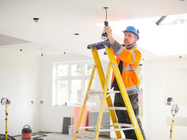 Worker in safety gear installing a light fixture on a ladder indoors.