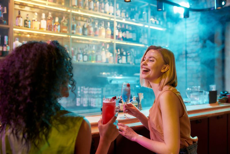 Moments of Happiness. Two women having fun, drinking cocktails while sitting at the bar counter. Friends spending time at night club, restaurant. Selective focus on woman. Horizontal shot