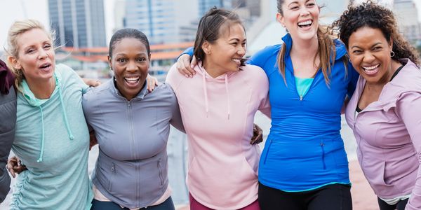 Five diverse women smiling and embracing outdoors in athletic wear.