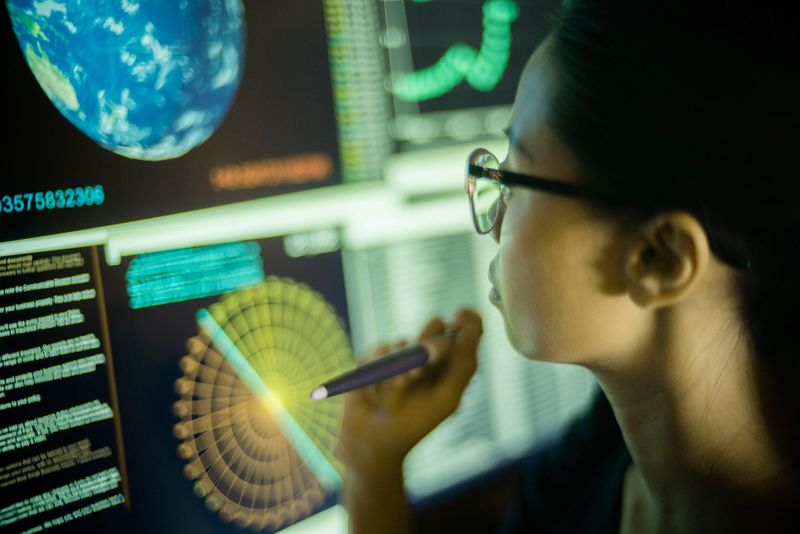 Stock photograph of a young Asian woman, pointing at, and learning from, global data displayed on a large computer monitor.
