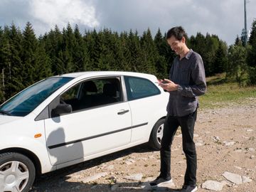 Man standing beside a white car on a dirt road, smiling at his phone.