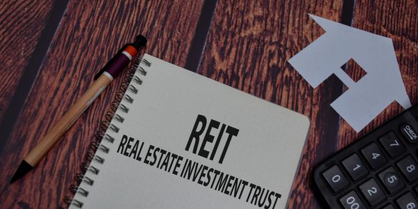 Notebook with 'REIT Real Estate Investment Trust' text, pen, calculator, and paper house on wooden surface.
