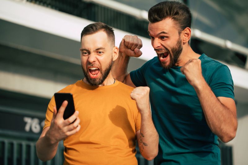 Two happy excited fan friends in euphoria mood after winning in a bet with a smartphone in hand on stadion background