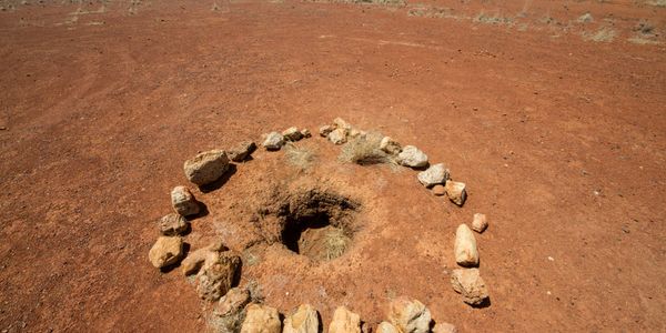 A hole in red desert soil surrounded by a circle of rocks.