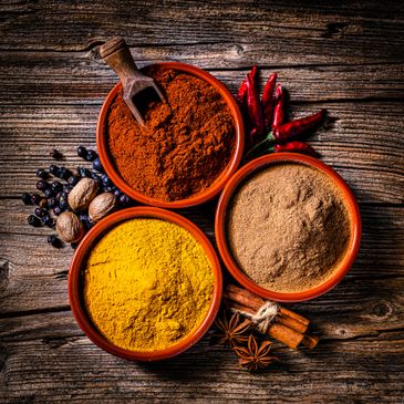 Three bowls of colorful spices on a rustic wooden table with whole spices.